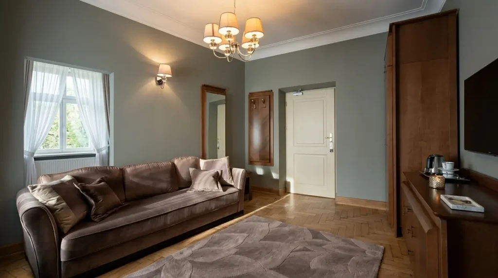 Szeroki kadr na luksusowy apartament w stylu pałacowym. W pokoju stoi duża, brązowa, pikowana sofa Chesterfield umieszczona na ozdobnym dywanie. Ściany są gładkie, w odcieniu stonowanej szarości, a w głębi widać wysokie, klasyczne drzwi wewnętrzne.